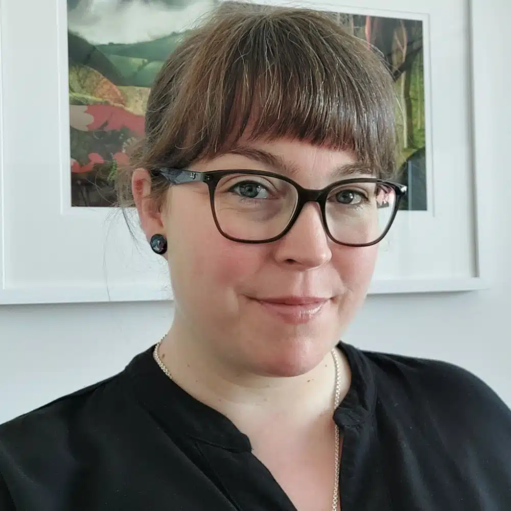 A woman with brown hair and fringe, wearing glasses, black earrings, and a black top, smiles at the camera. A framed nature artwork hangs on the white wall behind her—she's part of the Kilkenny Animated Festival Team.