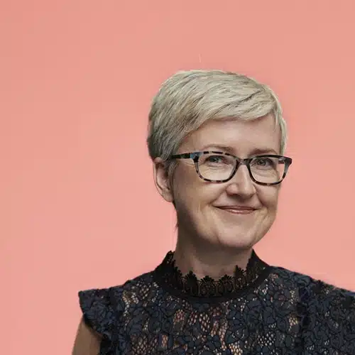 Smiling woman with short blonde hair and glasses, part of the Kilkenny Animated Festival Team, wears a dark lace top and stands in front of a solid peach-coloured background.