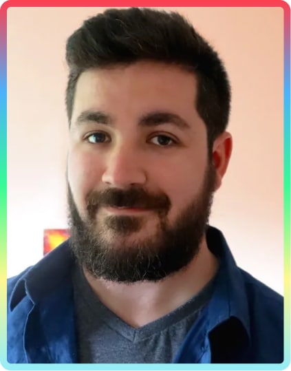 A man with short dark hair and a full beard smiles softly at the camera at the Kilkenny Animated Festival. He is wearing a dark blue shirt over a grey T-shirt. The background is softly lit with pastel colours and a rainbow-coloured border frames the image.