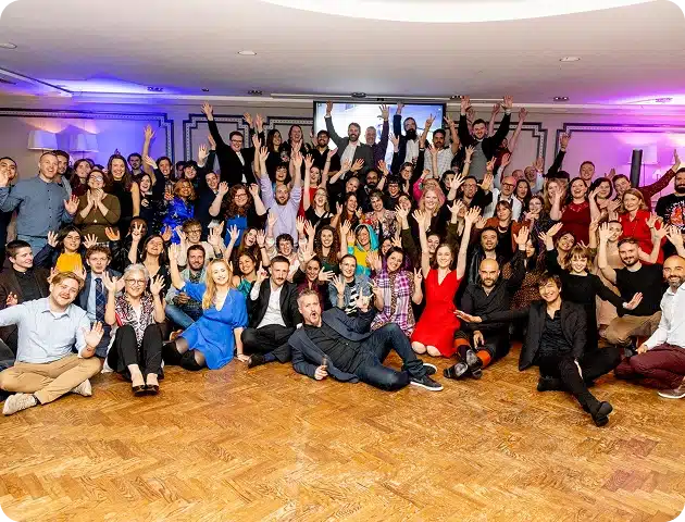 A large, diverse group of people pose together in a brightly lit room, smiling and waving at the camera. Some are standing, some sitting, dressed in semi-formal and casual attire, capturing the festive spirit of the Kilkenny Animated Festival.