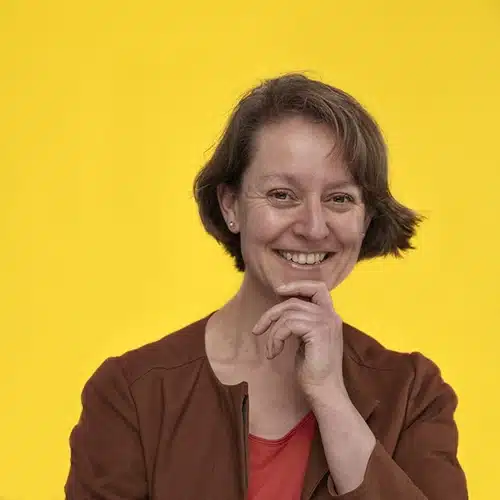 A woman with short brown hair smiles and rests her hand on her chin. She is wearing a rust-coloured jacket over a red top, standing in front of a bright yellow background, representing the Kilkenny Animated Festival Team.