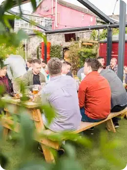 A group of people sits around a wooden picnic table outdoors, enjoying drinks and conversation in a lively garden setting during the Explore Kilkenny Animated Festival. Green grass, colourful buildings, and leafy branches frame the vibrant scene.
