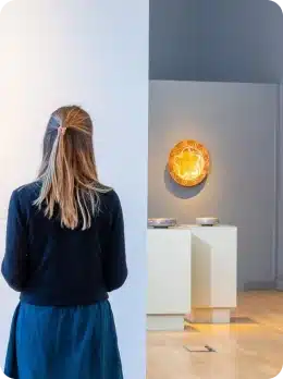 A woman with blonde hair, seen from behind, stands in an art gallery looking at illuminated artwork on a grey wall—an experience reminiscent of moments at Explore Kilkenny Animated Festival. Display plinths and plates appear in the background.