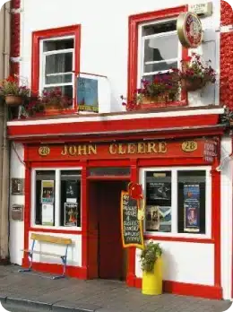 The image shows the front of a pub called "John Cleere", with red-trimmed windows and door, hanging flower pots, posters in the windows, and a yellow barrel with plants—perfect for those looking to explore Kilkenny Animated Festival on a city pavement.
