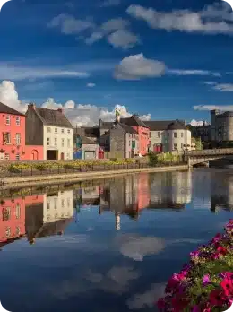 Colourful houses line a riverside under a bright blue sky with scattered clouds, their reflections visible in the calm water; vibrant flowers bloom in the foreground—perfect for those who wish to explore Kilkenny Animated Festival nearby.