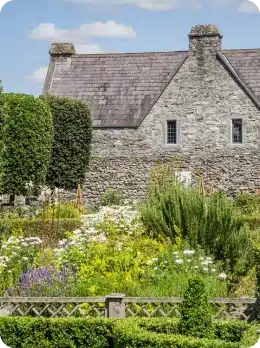 A stone cottage with a slate roof stands behind a lush, colourful garden filled with blooming flowers—an inviting scene to explore before heading to the Kilkenny Animated Festival under blue skies and scattered clouds.