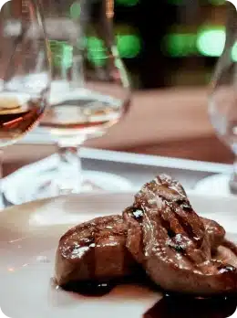 Two pieces of seared foie gras with sauce rest on a white plate, accompanied by two glasses of amber-coloured wine—a perfect pairing to enjoy as you explore Kilkenny Animated Festival in a vibrant, green-lit setting.