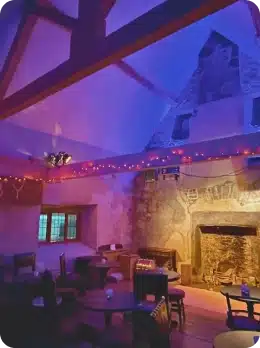 A cosy room with stone walls, wooden beams, and a large fireplace. Warm purple and orange lighting, fairy lights, and small round tables invite you to relax—an intimate spot to unwind after you explore Kilkenny Animated Festival.