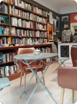A cosy café with bookshelves filled with books, brown chairs, and a small round table with a teacup and saucer—perfect for relaxing after you explore Kilkenny Animated Festival. A chalkboard menu and coffee counter are visible in the background.