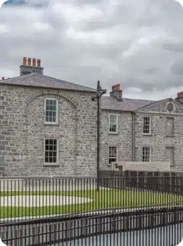 A stone building with arched and rectangular windows stands behind a black metal fence under a cloudy sky. Discover its charm as you explore Kilkenny Animated Festival, with a neatly trimmed lawn and a sign near the entrance welcoming visitors.