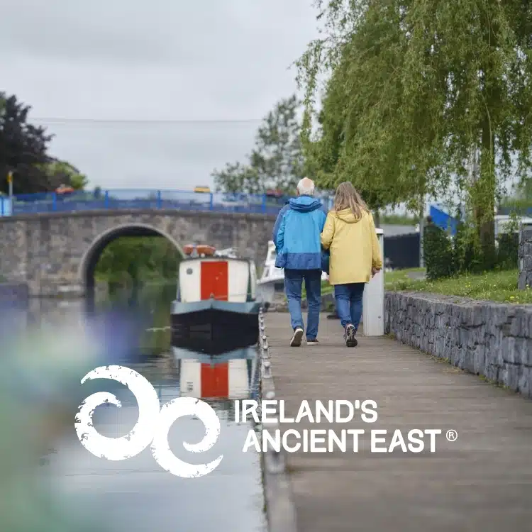 An older couple walks hand in hand along a canal towpath beside a calm waterway, with a stone bridge and moored boat in the background. The words "Explore Kilkenny Animated Festival," "Ireland's Ancient East," and a white logo are overlaid.