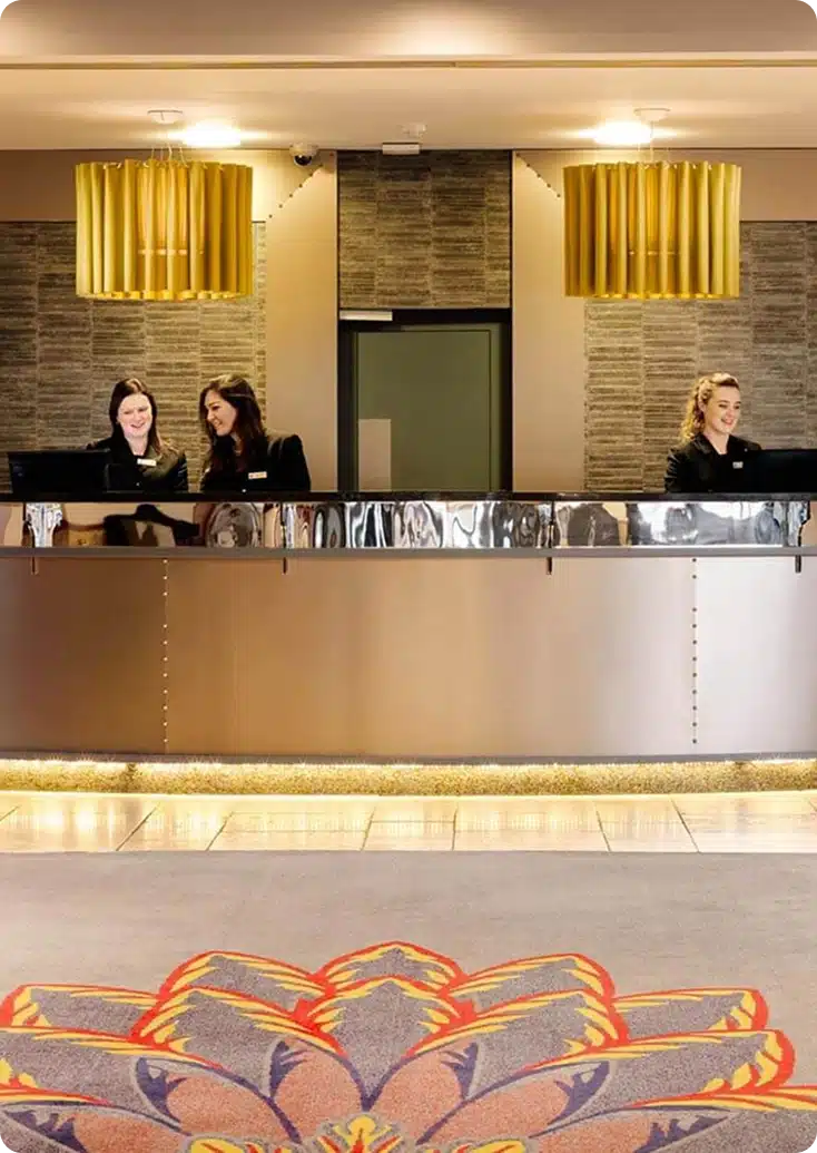 Three women in black uniforms stand behind a modern hotel reception desk with a metallic finish. The lobby, featuring two large gold hanging lights and textured wall panels, highlights the welcoming presence of Kilkenny Animated Festival Sponsors.