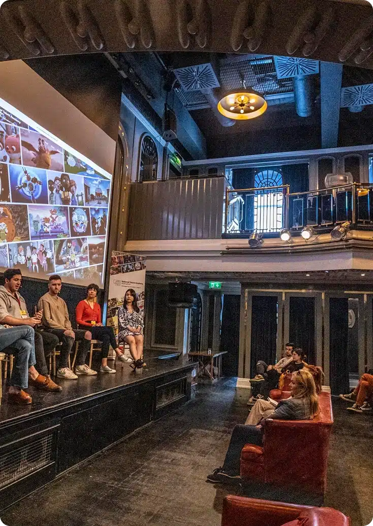A panel of four people sits on stage in front of a large screen displaying various images, as a small audience watches in a dimly lit, modern event space—capturing the vibrant atmosphere of About Kilkenny Animated Festival.