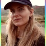 A woman with long blonde hair wearing a navy cap and black shirt stands outdoors with hills in the background, a colourful border framing her as if she's part of the Kilkenny Animated Festival. She has a neutral expression, small hoop earrings, and a necklace.