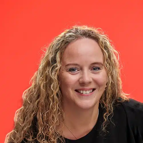A woman with long, curly blonde hair and a black top, part of the Kilkenny Animated Festival Team, smiles in front of a bright orange background.