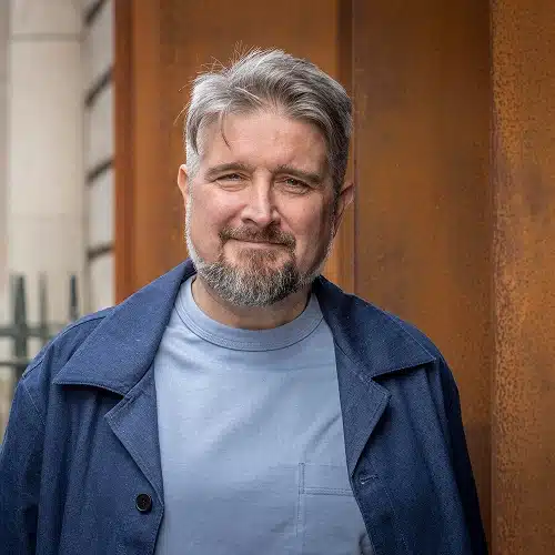 A middle-aged man with grey hair and a beard smiles at the camera. Wearing a light blue shirt and dark blue jacket, he stands outdoors by a rust-coloured wall and metal fence, representing the Kilkenny Animated Festival Team.