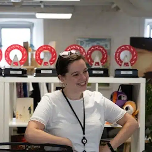 A member of the Kilkenny Animated Festival Team, wearing a white shirt and lanyard, stands smiling in front of a shelf with red and white circular awards and soft toys in a bright, modern room.