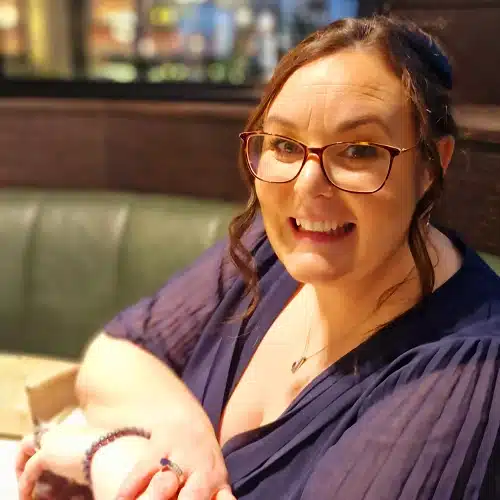 A woman with glasses and wavy brown hair, wearing a navy blue top, is smiling whilst sitting at a booth in a restaurant, possibly celebrating with the Kilkenny Animated Festival Team.