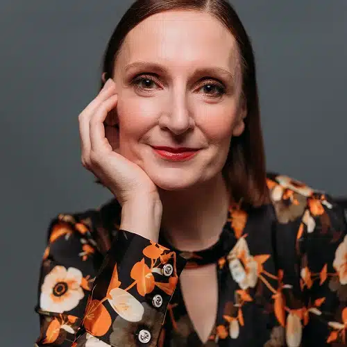 A woman with straight brown hair, wearing a black blouse with orange and white floral patterns, rests her head on her hand and smiles gently at the camera, representing the Kilkenny Animated Festival Team against a plain dark background.