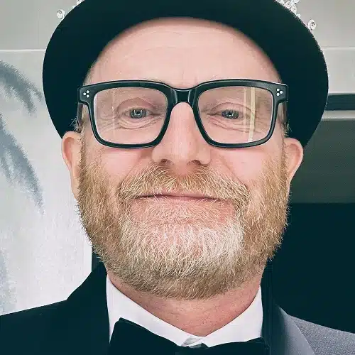 A middle-aged man with a reddish-blond beard and glasses smiles at the camera. He is wearing a black hat, a black dinner jacket, and a bow tie, representing the Kilkenny Animated Festival Team indoors against light-coloured walls.