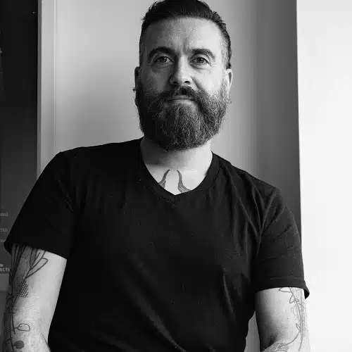 A man with a beard and tattoos sits by a window, wearing a black t-shirt. The image is in black and white, and he looks directly at the camera with a calm expression—evoking the creative energy of the Kilkenny Animated Festival Team.