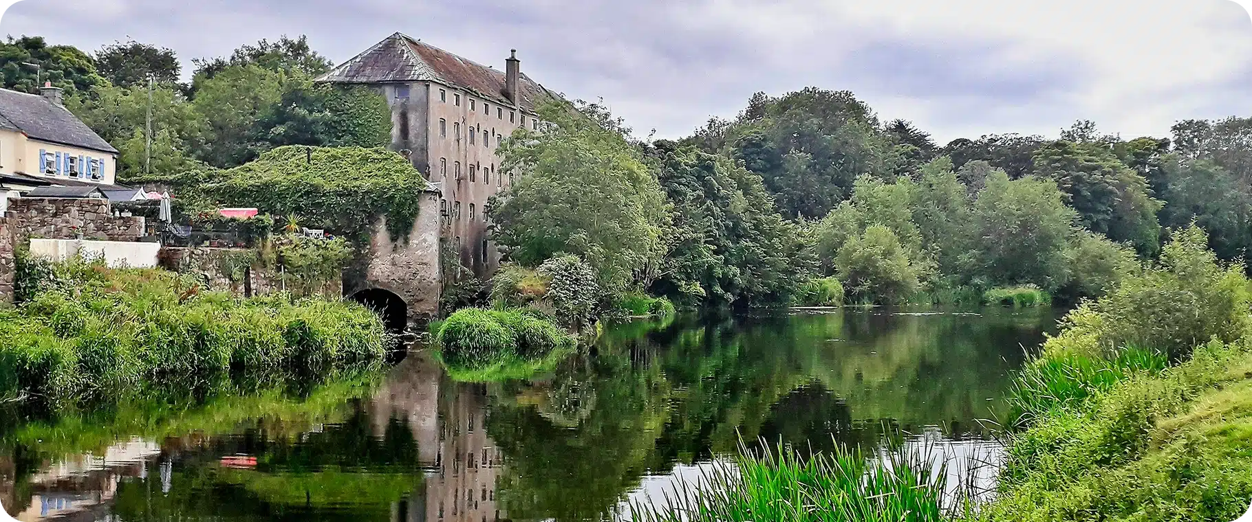 A historic stone building covered in ivy stands beside a calm river, surrounded by lush green trees and tall grass. Explore Kilkenny Animated Festival amidst reflections of this scenic view beneath a cloudy sky.