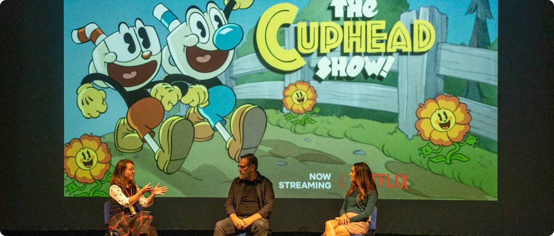 Three people sit and talk onstage in front of a large screen displaying colourful artwork and the title "The Cuphead Show!" at the Kilkenny Animated Festival, with animated characters, flowers, and a "Now Streaming on Netflix" message.