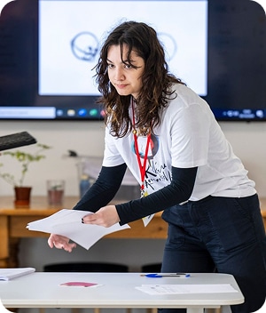 A woman with wavy hair wearing a white T-shirt and red lanyard stands at a table, sorting papers during the Kilkenny Animated Festival. A screen with graphics is visible behind her in a classroom or office setting.