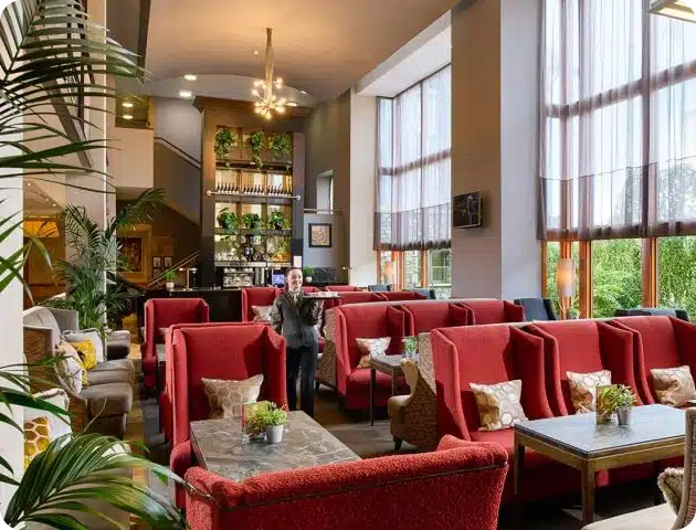 A modern lounge with tall windows, red high-backed chairs, small tables with potted plants, and a barman by the bar—an inviting space, often enjoyed by Kilkenny Animated Festival Sponsors, and bathed in natural light.