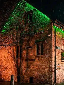 A stone building at night is illuminated by green and orange lights. The sign "Miller's Inn" glows above the door, raindrops falling in the dark. Trees partially frame the scene—an inviting stop as you explore Kilkenny Animated Festival.