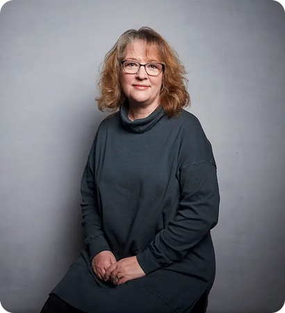A woman with shoulder-length curly hair and glasses sits on a stool against a plain grey background, wearing a dark long-sleeve polo-neck tunic and smiling gently at the camera, embodying the creative spirit of About Kilkenny Animated Festival.