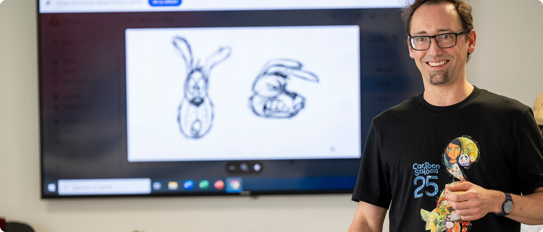 A man wearing glasses and a Cartoon Saloon 25th anniversary T-shirt smiles in front of a screen showing two cartoon bunny sketches, one friendly and one angry, at an event about Kilkenny Animated Festival.