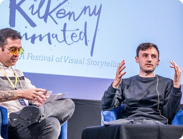 Two men sit on stage at the Kilkenny Animated Festival in front of a screen displaying "A Festival of Visual Storytelling." One holds papers while the other speaks and gestures, both dressed in casual clothing.