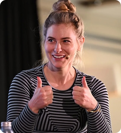 A woman with blonde hair in a bun, wearing a black and white striped shirt and a headset microphone, is smiling and giving two thumbs up, ready to share About Kilkenny Animated Festival.