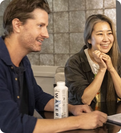 Two people sit at a table smiling and talking during a meeting about Kilkenny Animated Festival. A carton of water and some papers are on the table in front of them. The background features a textured wall.