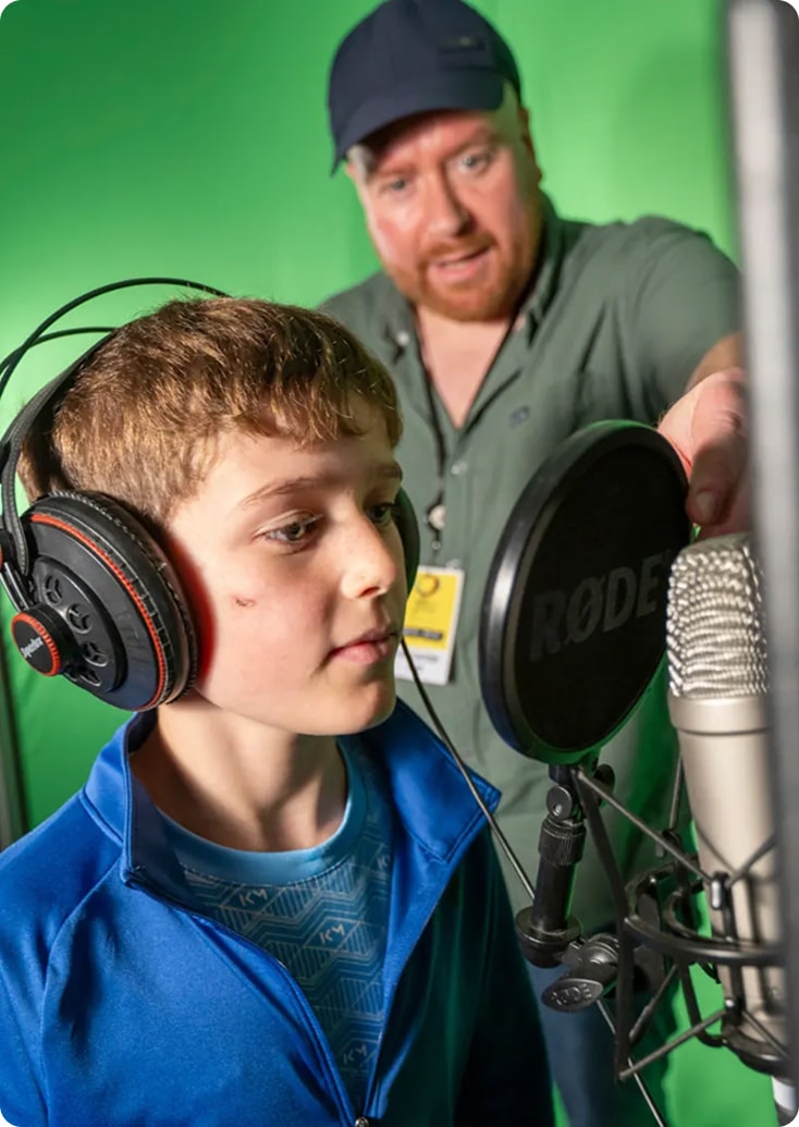 A boy wearing headphones stands in front of a microphone, speaking or singing, as a bearded man in a cap adjusts the mic in a studio with a green screen background at the Kilkenny Animated Festival.