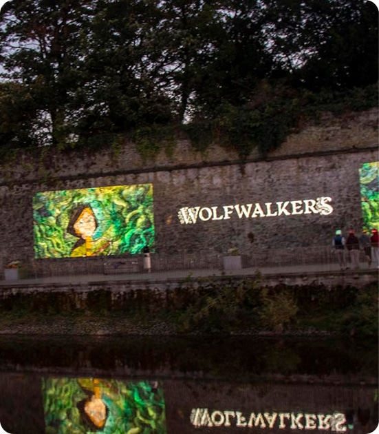 A stone wall at dusk displays illuminated "WOLFWALKERS" text and a colourful mural of a girl in a green forest, reflected in the water below—an enchanting scene from About Kilkenny Animated Festival, as several people walk along the path nearby.