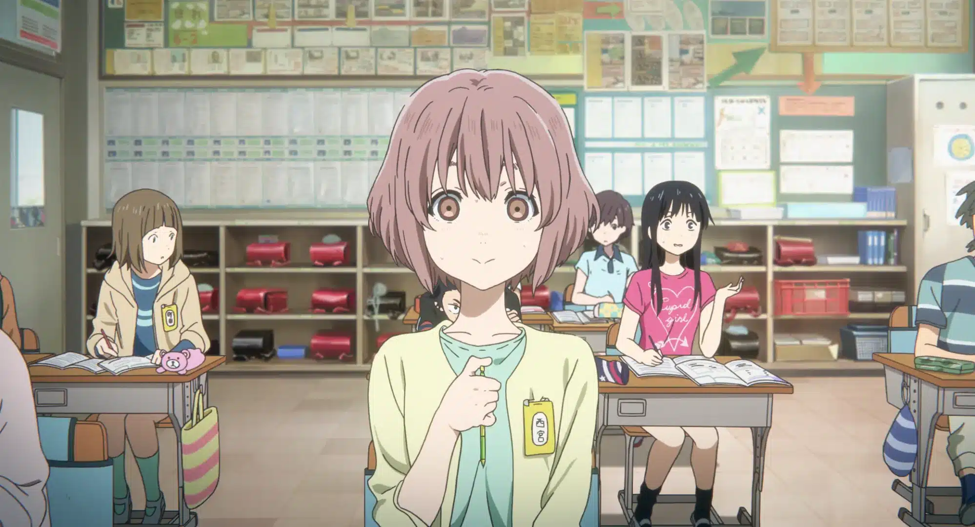 A girl with short brown hair stands in a colourful classroom, holding her shirt collar. Other pupils sit at desks behind her, studying and chatting. The classroom walls are covered with posters and charts.