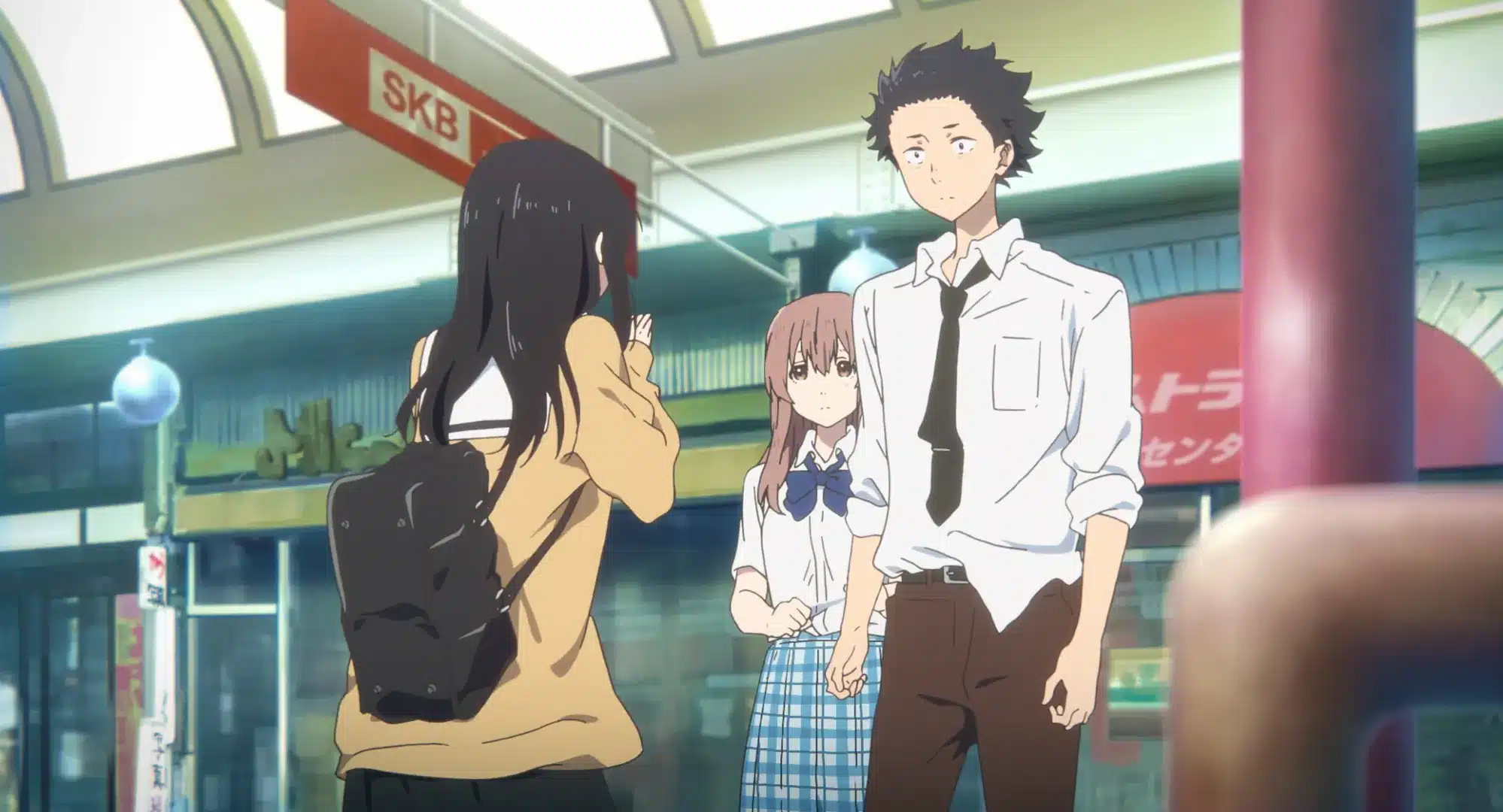 A girl with long black hair and a rucksack faces a boy and a girl in school uniforms standing outside a shop in a covered shopping street.