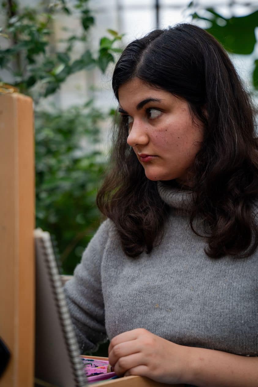 A woman with long dark hair and a grey jumper looks to the side while drawing in a sketchbook, surrounded by green plants.