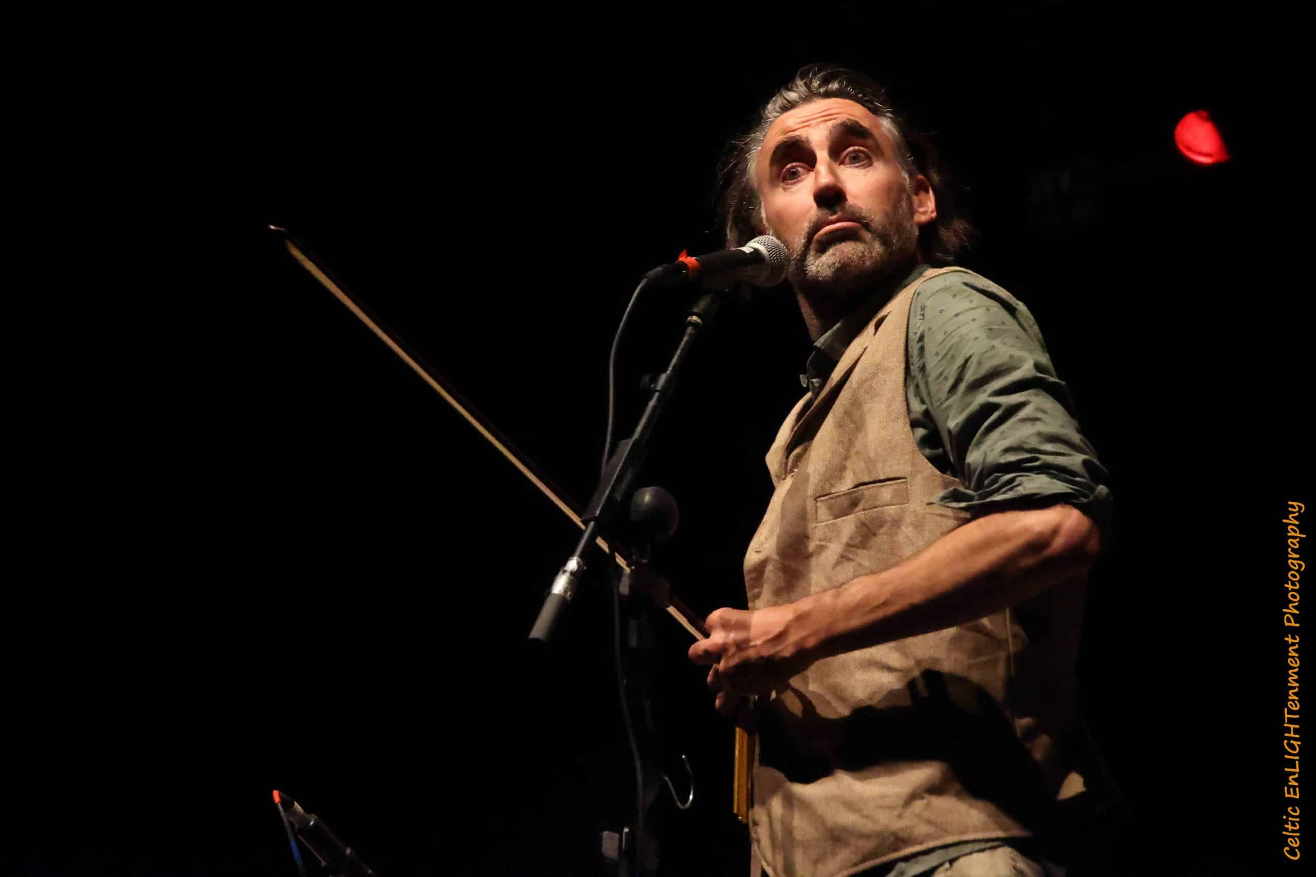 A man with long hair and a beard plays a stringed instrument with a bow on stage, looking surprised. He stands by a microphone, wearing a waistcoat over a shirt, with dramatic lighting highlighting his face.
