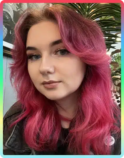 A person with long, wavy pink hair and light make-up looks at the camera. They are indoors with green plants and framed art in the background—a scene reminiscent of creative gatherings like Kilkenny Animated Festival. The photo has a subtle coloured border.