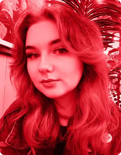 A person with wavy hair looks at the camera while sitting indoors, with leafy plants in the background. The image, reminiscent of creative moments at Kilkenny Animated Festival, has a strong red filter.