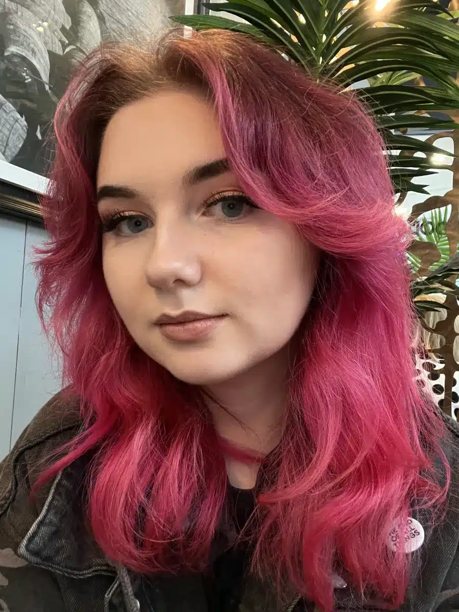 A person with wavy, bright pink hair and fair skin looks at the camera, sitting indoors with leafy plants and framed artwork visible in the background.