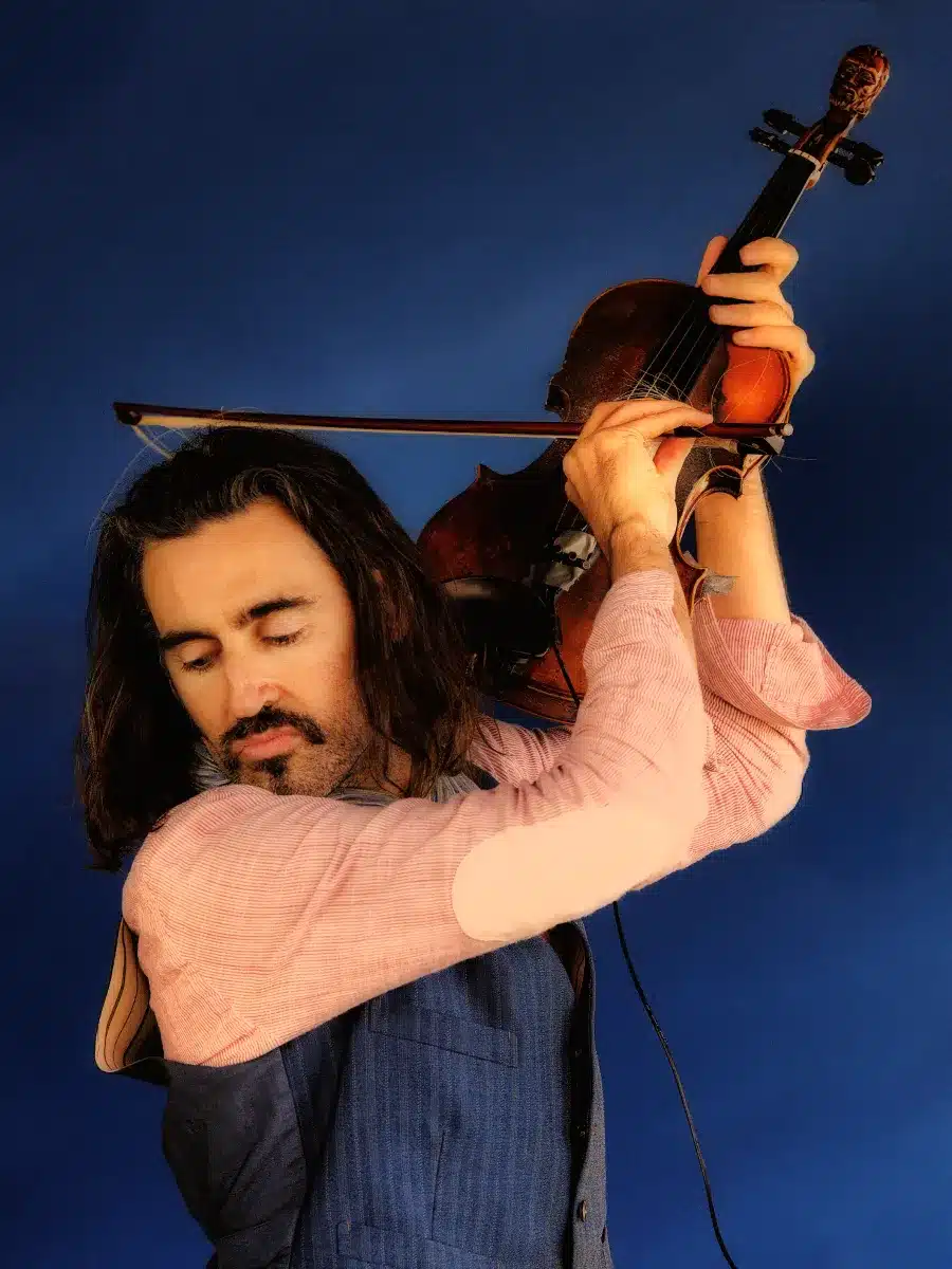 A man with long brown hair and a beard plays a violin passionately, eyes closed, wearing a pink shirt and blue waistcoat against a solid blue background.