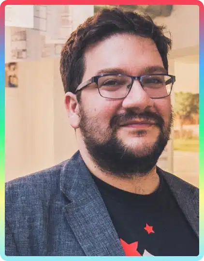 A man with short dark hair, glasses, and a beard wears a grey jacket over a black shirt with red and white designs. He is smiling and standing indoors at the Kilkenny Animated Festival, with blurred artwork in the background.