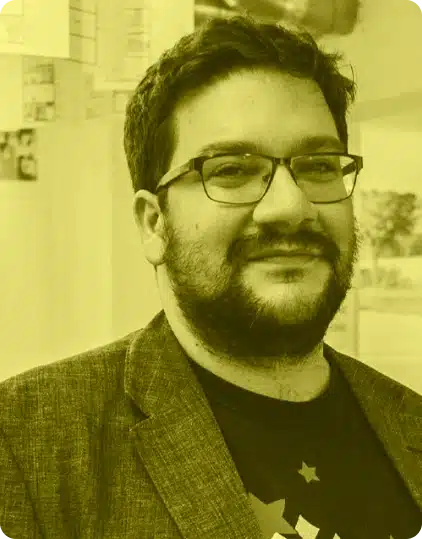 A man with dark hair, glasses, and a beard is smiling slightly. He is wearing a blazer over a T-shirt. The image, taken at the Kilkenny Animated Festival, has a yellow tint and a blurred indoor background.