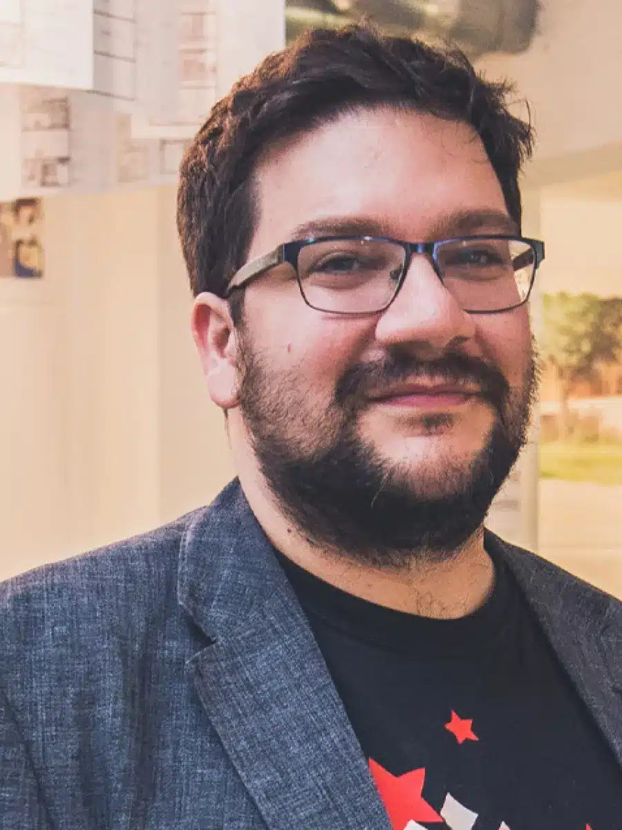 A bearded man with glasses wearing a grey blazer and black T-shirt smiles whilst standing indoors with a modern background.
