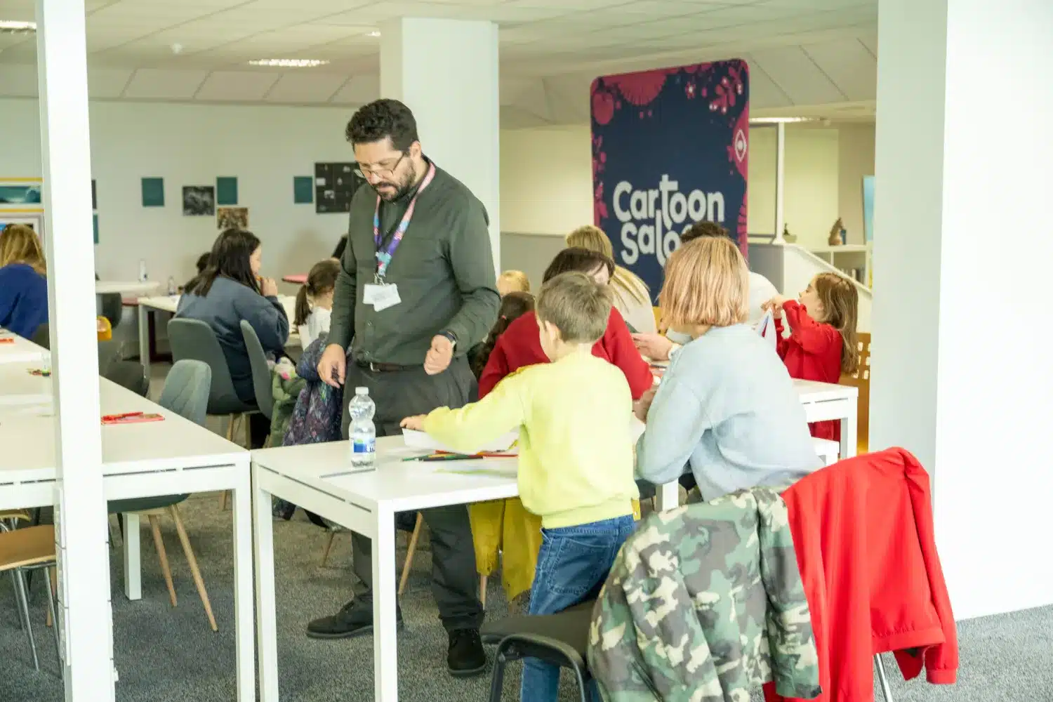 A group of children and adults sit and stand around tables, drawing and colouring in a brightly lit room with a “Cartoon Salon” sign on the wall.