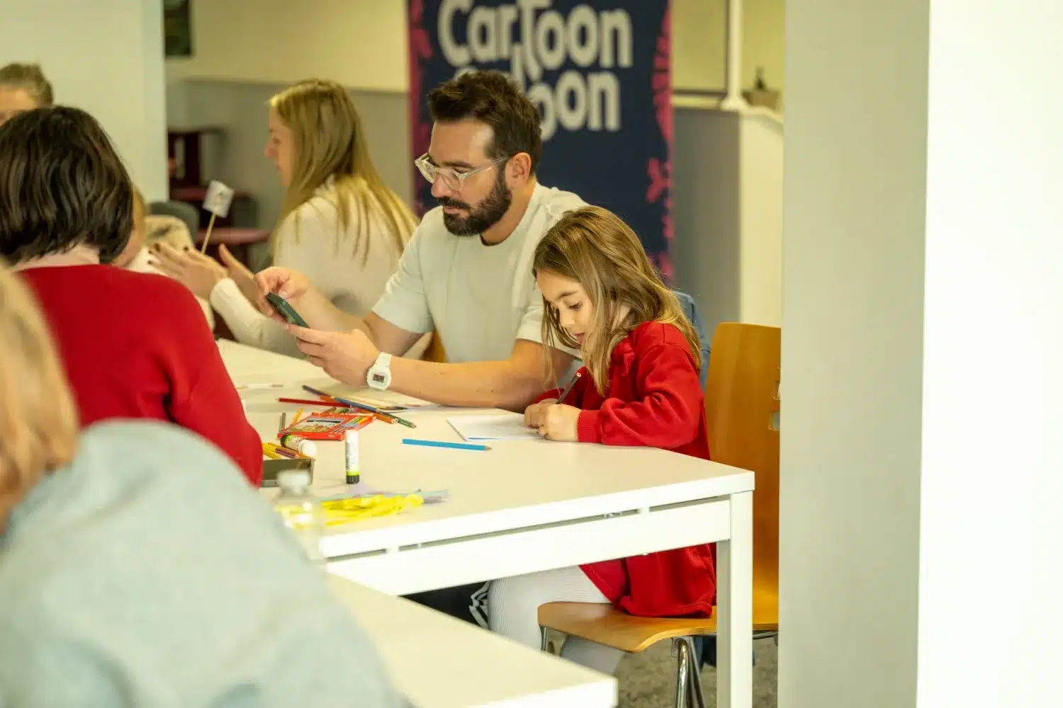 A man and a young girl sit at a table, drawing with coloured pencils. Other people are also seated around the table. A "Cartoon" sign is visible in the background. The atmosphere appears creative and focused.